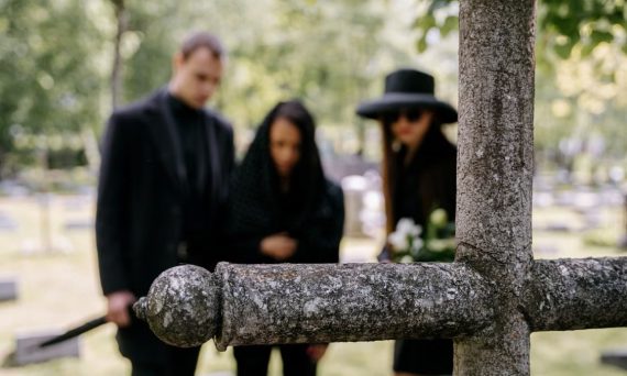 A family gathers in grief around a cross monument in a peaceful cemetery setting.